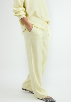 Osoba oblečená v voľné bledozelené nohavice a zodpovedajúcu košeľu, ruky v kapsách, stojaca s jednou nohou mierne vpredu v sivých balerínach.