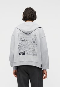 Άτομο που φοράει ανοιχτό γκρι φούτερ με κουκούλα, με μαύρα γραφικά σχέδια σε στυλ χειρόγραφου στο πίσω μέρος, συνδυασμένο με σκούρο παντελόνι, στραμμένο με την πλάτη προς τον θεατή.