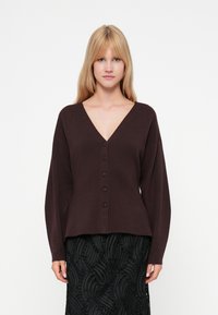 Cardigan marrone scuro con scollo a V, maniche lunghe e cinque bottoni frontali. Texture morbida e vestibilità rilassata, abbinato a una gonna nera strutturata.