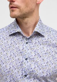 Camicia con bottoni dal motivo floreale in viola, blu e verde su uno sfondo chiaro. Presenta un colletto classico e bottoni scuri.