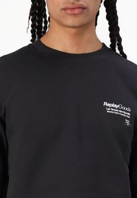 Felpa nera con collo a costine; presenta il logo testuale bianco "ReplayGoods" e altre lettere. Tessuto liscio e di peso medio.