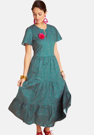 Woman wearing a teal polka dot midi dress with flutter sleeves, pink flower brooch, pink earrings, gold bangles, and red shoes, holding the skirt.