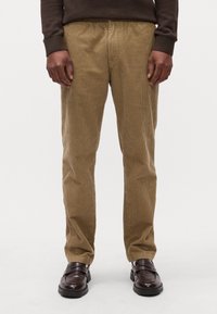 Pantalons en velours côtelé de teinte marron clair avec une texture à côtes verticales ; associés à des chaussures décontractées marron foncé. Détail de la taille élastique visible.