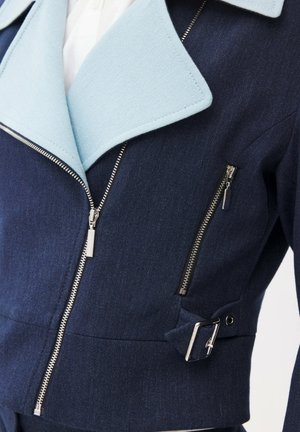 Marineblå jakke med lyseblå revers, sølv lynlåse foran og på brystet, samt en sølv spænde detalje på siden.