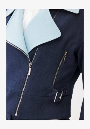 Marineblå jakke med lyseblå revers, sølv lynlåse foran og på brystet, samt en sølv spænde detalje på siden.