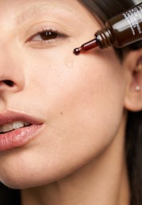 Brun dropperflaska som dispenserar ett klart serum på huden, som visar en jämn textur och naturlig hy. Närbild på ansiktsdrag.