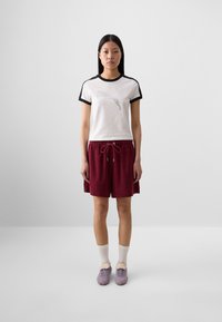 A young woman stands in front of a neutral background, wearing a white Puma T-shirt, burgundy shorts, white socks, and purple sneakers.
