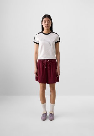 A young woman stands in front of a neutral background, wearing a white Puma T-shirt, burgundy shorts, white socks, and purple sneakers.