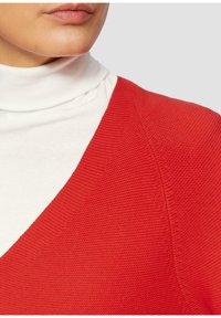 Rode textuurtrui met een V-hals ontwerp, gelaagd over een witte coltrui. Glad materiaal met zichtbare stiksels.