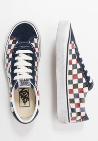 Pari lippuja Vans, joissa on punainen, sininen, musta ja valkoinen ruudullinen kuvio, valkoiset nauhat sekä tummansininen mokkanahka kärjessä ja kannassa.