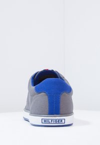 Šedé tenisky s modrým detailem na patě a bílou podrážkou, na zadní části s označením "HILFIGER", umístěné na neutrálním bílém pozadí.