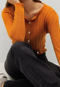 Pomarańczowy kardigan dzianinowy z dodatkiem warkocza, z okrągłymi, srebrnymi guzikami i ściągaczami, zestawiony z ciemnymi spodniami i szarymi butami.