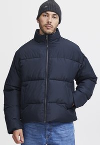 Navy puffer jacket with high collar, front zip, and quilted design. Gray knit beanie worn. Casual fit with side pockets and structured shoulders.