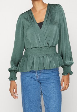 Femme portant un chemisier croisé vert à manches longues avec une taille et des poignets smockés, associé à un jean bleu taille haute.