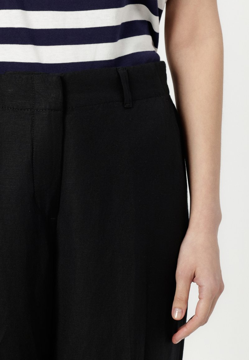 Czarne lniane spodnie z płaskim pasem i subtelnymi bocznymi kieszeniami. Połączone z granatowo-białą, pasiastą bluzką. Gładka tekstura i luźny krój.