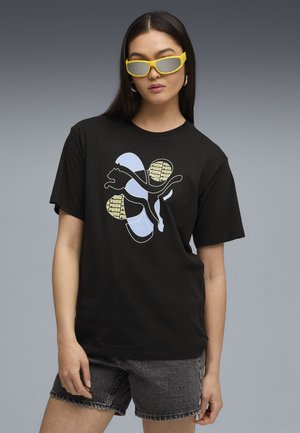 Un tricou negru din bumbac cu un logo Puma stilizat în nuanțe de albastru deschis și accente galbene. Modelul poartă pantaloni scurți din denim gri și ochelari de soare galbeni.