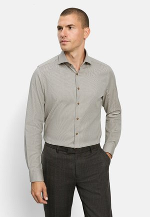 Uomo con i capelli corti che indossa una camicia grigio chiaro a maniche lunghe con bottoni e pantaloni grigio scuro a quadri, in piedi con una mano in tasca.
