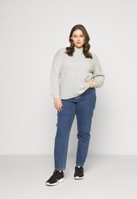 Suéter cinza de malha com gola alta, combinado com jeans de denim azuis e tênis atléticos pretos. O suéter tem um acabamento texturizado.