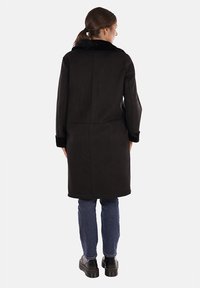 Cappotto in finta suede nero con un morbido colletto in pelliccia sintetica, maniche lunghe e design lungo fino al ginocchio. Le cuciture visibili e le tasche aggiungono un dettaglio sottile.