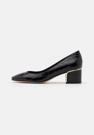 Escarpin en cuir noir à motif crocodile avec un petit talon carré souligné d'une fine ligne dorée, bout pointu et semelle beige sur fond blanc.