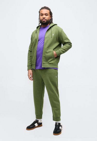 Άντρας με κοτσίδες dreadlocks που φοράει πράσινο φούτερ με φερμουάρ και παντελόνι φόρμας, μωβ μπλούζα, λευκές κάλτσες και μαύρα αθλητικά παπούτσια, στέκεται πάνω σε λευκό φόντο.