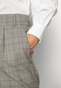 Grå rutiga byxor med hög midja, med veck och sidofickor, kombinerat med en texturerad vit långärmad skjorta.