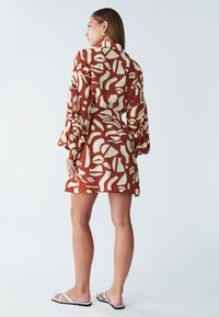 Sukienka o krótkich rękawach, długości do kolan, w kolorze rdzy z białymi abstrakcyjnymi wzorami; ma kołnierzyk i luźne rękawy, wykonana z lekkiego materiału.