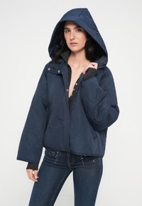 Μπουφάν navy μπλε με καπιτονέ επένδυση και κουκούλα, με κούμπωμα με πάτ και χαλαρές ραφές στους ώμους, σε κουτί σχήμα. Φορεμένο πάνω από μαύρη μπλούζα και τζιν.