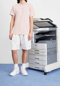 Ljusrosa bomullströja, vita shorts och vita sneakers, stående bredvid en grå multifunktionsskrivare på en blå matta.