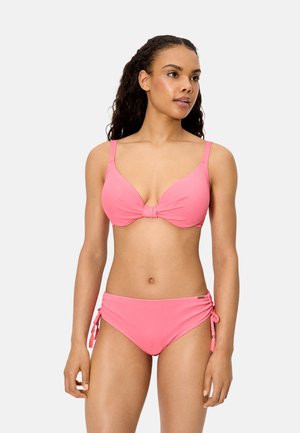 Mujer con cabello rizado que lleva un bikini rosa con un top anudado al frente y braguitas con lazos ajustables en los costados, posando frente a un fondo blanco liso.