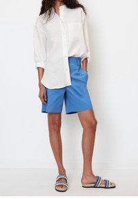 Hvid hørskjorte med knapper og frontlomme, kombineret med blå shorts. Fodtøjet har flettede stropper i marineblå og hvid.