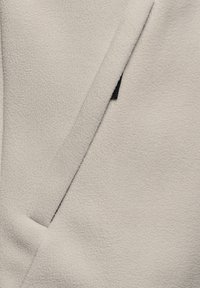 Ljust beige fleecejacka med en texturerad yta, omslagsdesign och en subtil svart accent vid fickområdet.