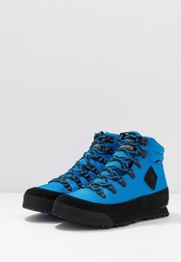 Vysoké turistické boty v jasně modré tkanině s černými gumovými podrážkami. Mají černé detaily a vzorované tkaničky pro pevné uchycení.