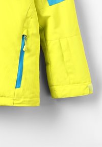 Ljusgul jacka med slät yta, med en blå dragkedjeficka och blå tygdetalj på axeln. Justerbara ärmslut.