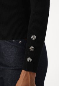 Pull noir côtelé avec des boutons argentés sur le poignet. Gros plan montrant la texture et les détails. Porté avec un jean foncé.