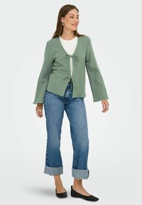 Cárdigan verde claro con lazo en el frente, hecho de tejido texturizado. Combinado con una camiseta blanca y jeans azules con puños enrollados, y zapatos planos negros.