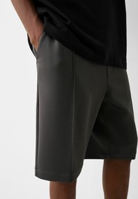 Shorts gris foncé en matériau texturé, avec une coupe au genou et un ourlet cousu. Des poches sont visibles sur les côtés.