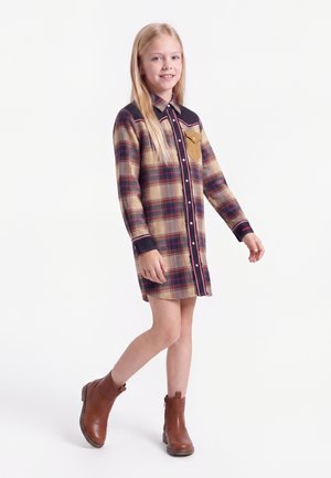 Pige med langt blondt hår iført en beige og rød ternet kjole og brune støvler til anklen, der går med et let smil mod en hvid baggrund.