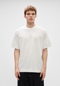 Λευκό βαμβακερό t-shirt με στρογγυλή λαιμόκοψη, κοντά μανίκια και ανάγλυφο λογότυπο στο κέντρο. Φοριέται με σκούρες τζιν παντελόνες.