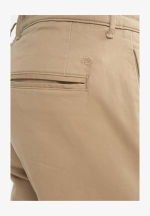 Beez värvi puuvillased püksid, millel on tagakott horisontaalse avaga ja diskreetne embroideeritud logo detail. Pehme tekstuur ja sile viimistlus.