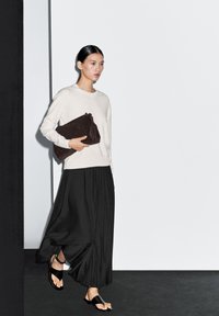 Lichtgrijze gebreide trui, zwarte plissé maxi rok en zwarte sandalen met bandjes. Het model draagt een donkerbruin suède clutch tasje.