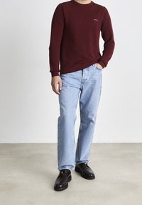 Bordeaux trui, blauwe spijkerbroek en donkerbruine schoenen. De trui heeft een geribbelde textuur en een klein logo op de borst. Casual pasvorm en ontwerp.
