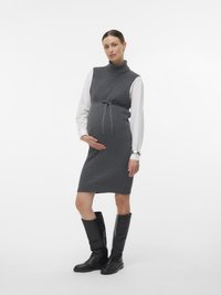 Kobieta w ciąży w szarej, bezrękawnikowej sukience dzianinowej na białej koszuli, czarnych, wysokich do kolan botkach, stojąca z jedną ręką na brzuchu, na neutralnym tle.