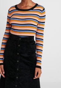 Pull à manches longues côtelé multicolore avec des rayures orange, bleu et noir, associé à une jupe en velours côtelé noire avec des détails de boutons.