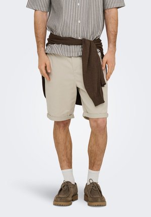 Homme portant un short beige retroussé, un pull marron noué autour de la taille, des chaussettes blanches et des chaussures en daim marron, debout sur un fond uni.