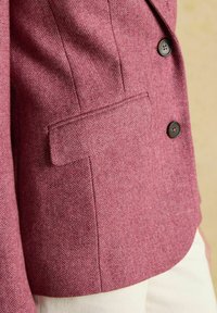 Detailný záber na ružový blazer so vzorom rybej kosti, s dvoma tmavohnedými gombíkmi a klopovým vreckom na bielych nohaviciach.