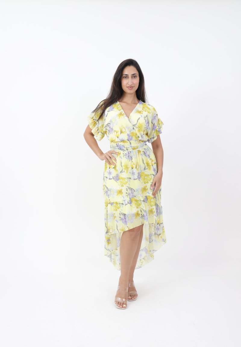 Gelbes florales Wickelkleid mit Rüschensäumen, asymmetrischem Saum und transparentem, geschichtetem Stoff; kombiniert mit klaren Sandalen mit Riemchen.