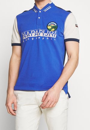 Mand, der bærer en blå og cremefarvet kortærmet polo-shirt med logo-tekst og et rundt mærke på brystet, parret med lyse bukser.