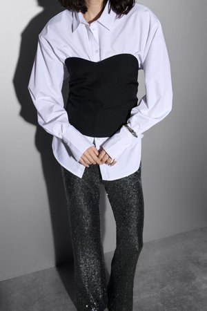 Weißes Button-Up-Hemd, das über einem schwarzen trägerlosen Top getragen wird, kombiniert mit figurbetonten, glitzernden schwarzen Hosen. Strukturierten Oberflächen und kontrastierenden Farben sind hervorstechend.