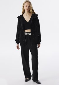 Felpa nera con zip decorata con borchie argento, abbinata a un top nero cropped e pantaloni larghi neri con un cinturino in vita con logo visibile.